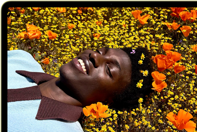 MacBook Neo, kleur Citrus, op het scherm een lachende persoon die in een zonnig bloemenveld ligt